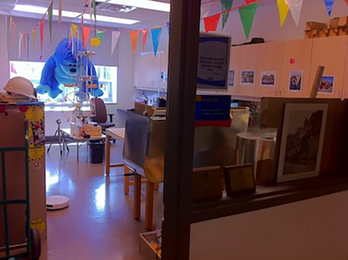 image of thrill lab, looking through glass window, large stuffed animal sits in window, flags hang from ceiling. black desktops and light wood cabinets line the walls
