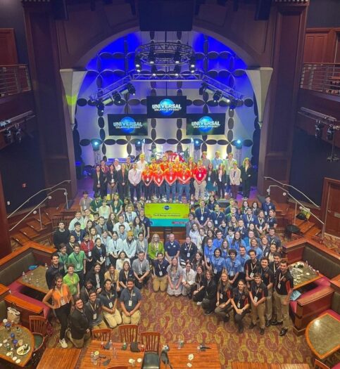 Photo shows TMTDI competitors at Universal's Rising Star venue standing on stage and in front of the stage. Some teams are clustered together wearing team polos. A large monitor displaying a competition background slide appears in the middle of the group.